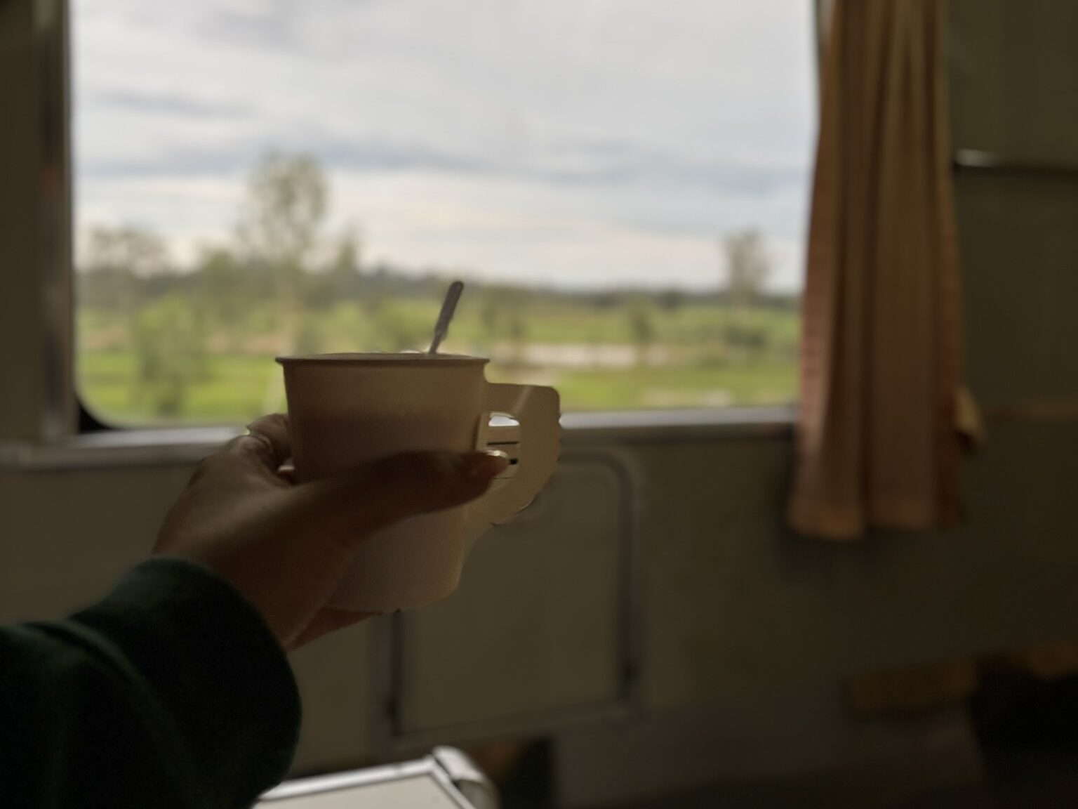 Hand holding a paper cup of coffee on the Bangkok to Vientiane overnight train with green fields visible through the window